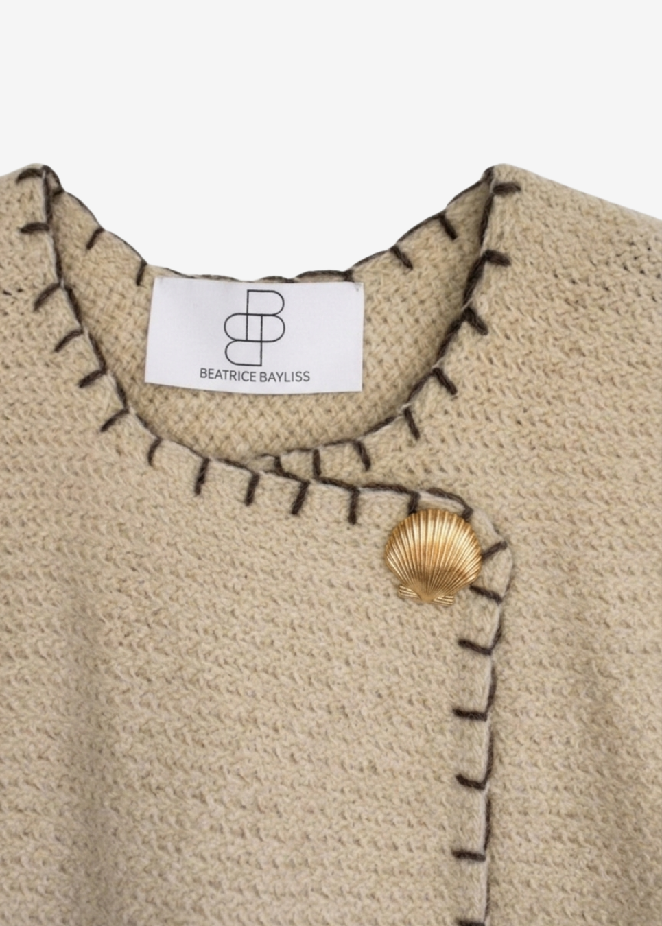 Close-up of a beige textured knit top by Beatrice Bayliss, featuring dark contrast stitching, a shell-shaped gold button, and a white brand label.