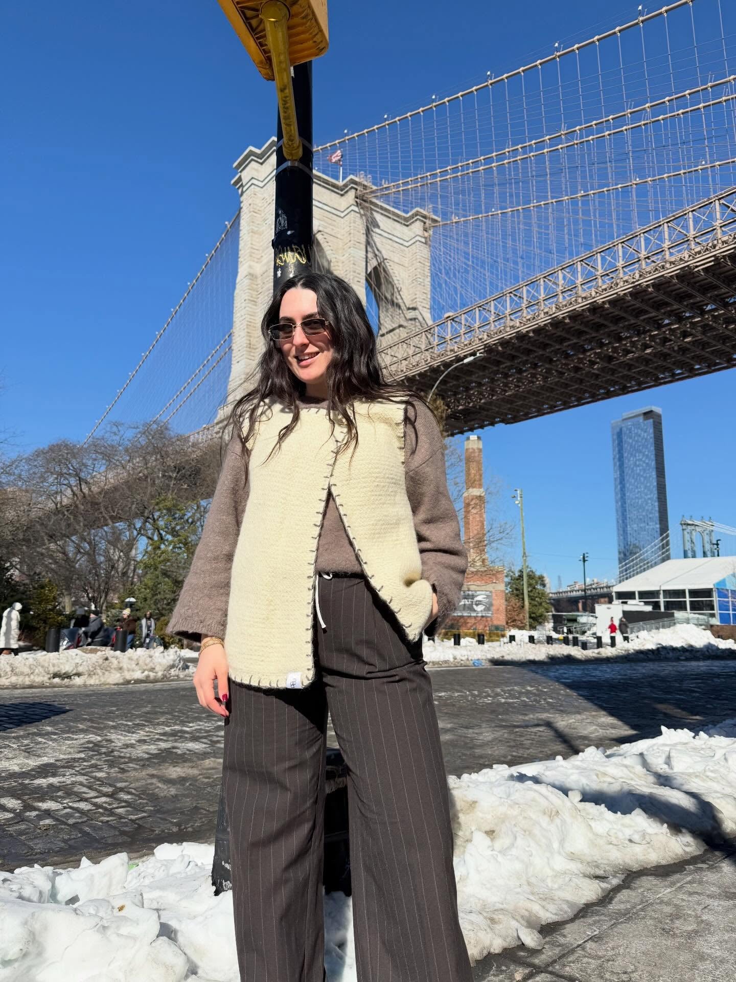 A woman wearing a cream cotswold wool-lined brown jacket and dark brown pinstripe trousers stands in front of the Brooklyn Bridge with snow on the ground and a clear blue sky.