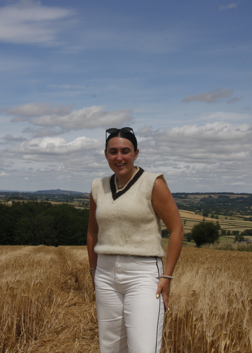 A woman standing in a golden wheat field under a blue sky with clouds, wearing a cream knit sleeveless sweater vest with black trim and white trousers.