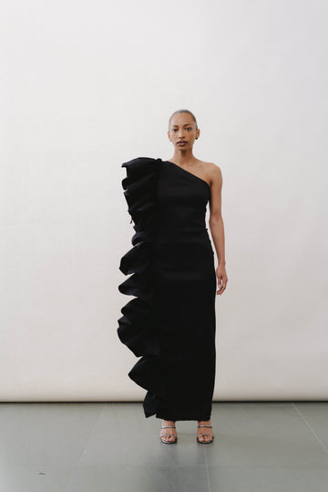 A model wearing a black asymmetrical tight-to-the-body taffeta dress with volume detail on the left side, standing against a white background.