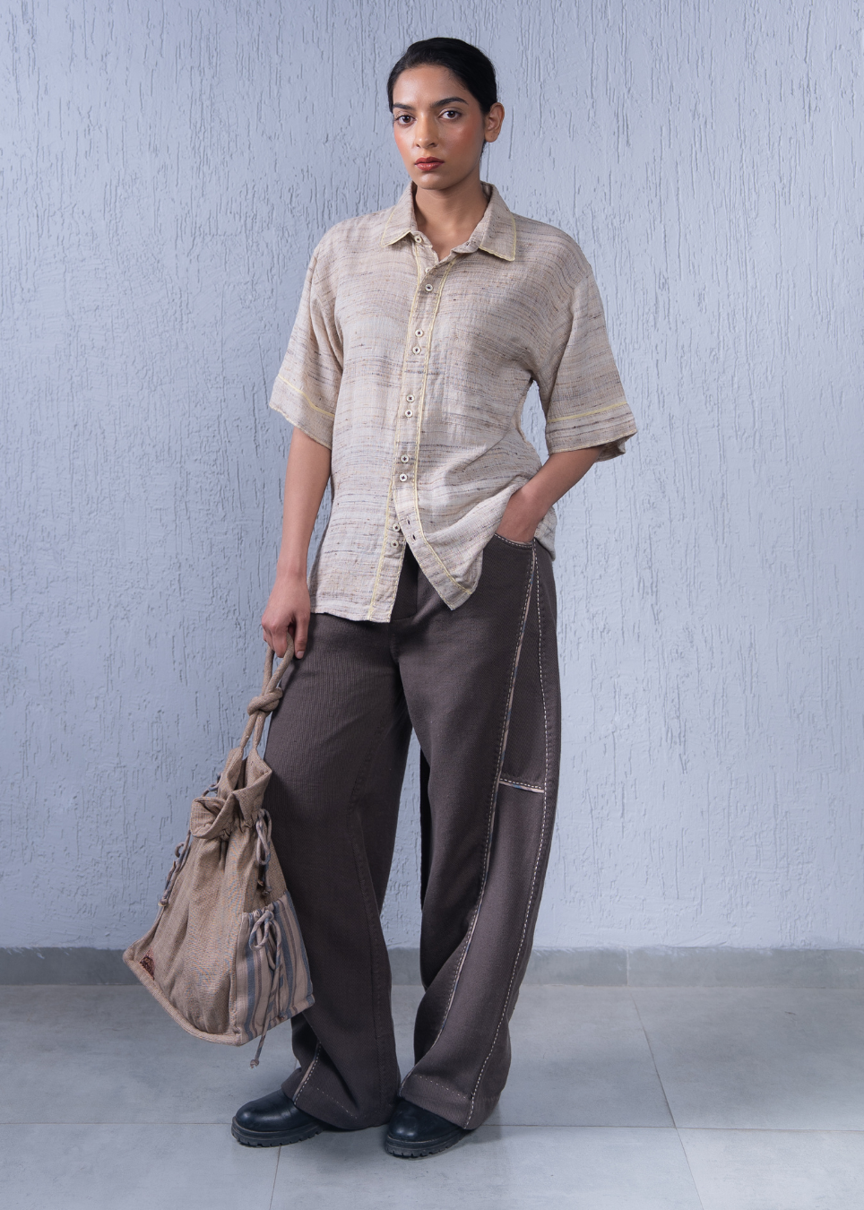A model stands against a textured light gray wall wearing a beige short-sleeve button-up shirt and dark brown wide-leg trousers, holding a beige canvas shoulder bag.
