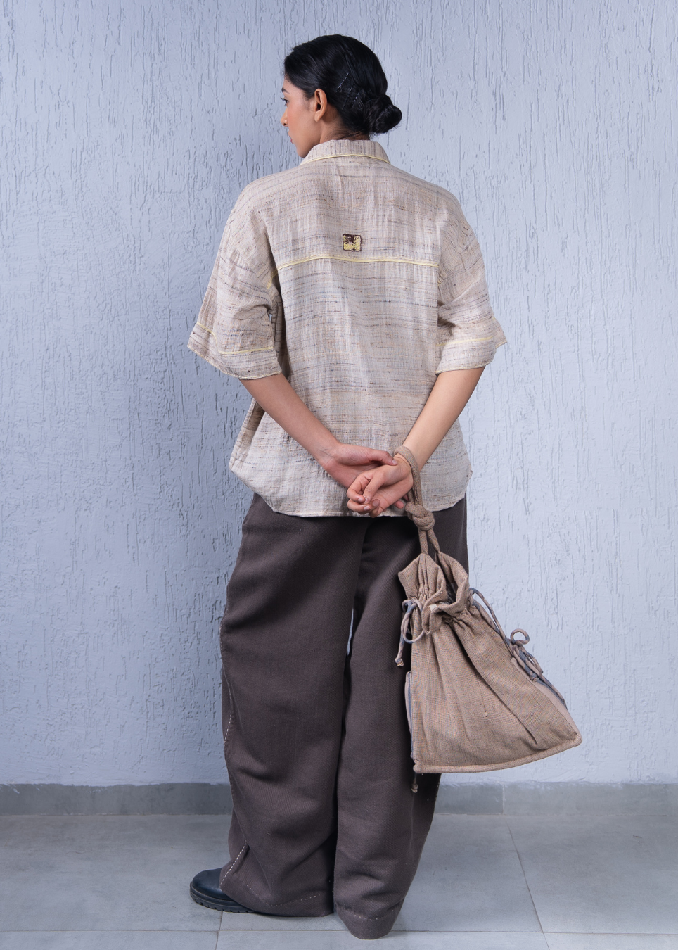 A woman stands with her back to the camera wearing a light beige linen shirt with a small embroidered patch on the upper back, dark brown wide-leg pants, and holding a beige drawstring bag against a textured light gray wall.