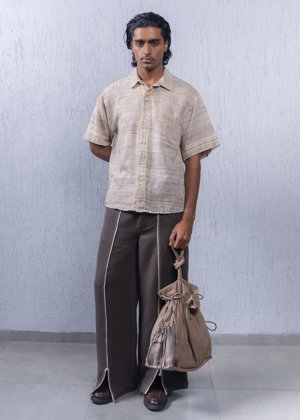 A man stands against a light gray textured wall wearing a beige short-sleeve button-up shirt, dark brown wide-leg trousers with white piping, and brown shoes, while holding a brown drawstring bag.