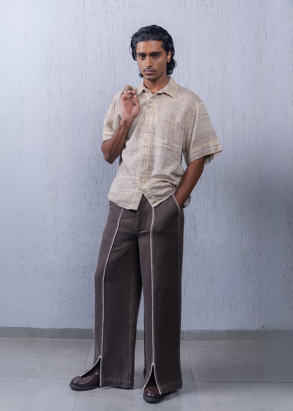 A man modeling a light beige short-sleeve linen button-up shirt and dark brown wide-leg trousers with white piping details, standing against a textured light gray wall.