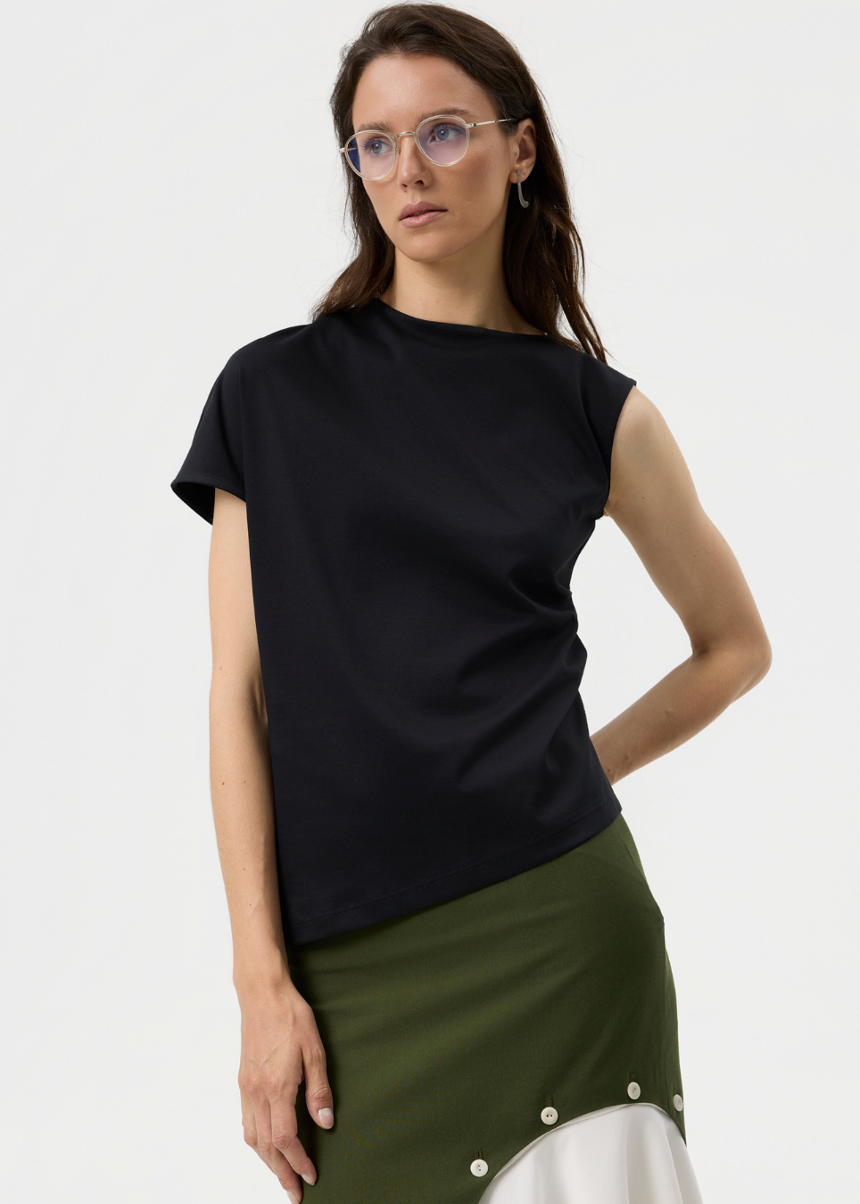 A woman wearing a black short-sleeve top and an olive green skirt with white buttons, photographed against a clean light gray studio background.