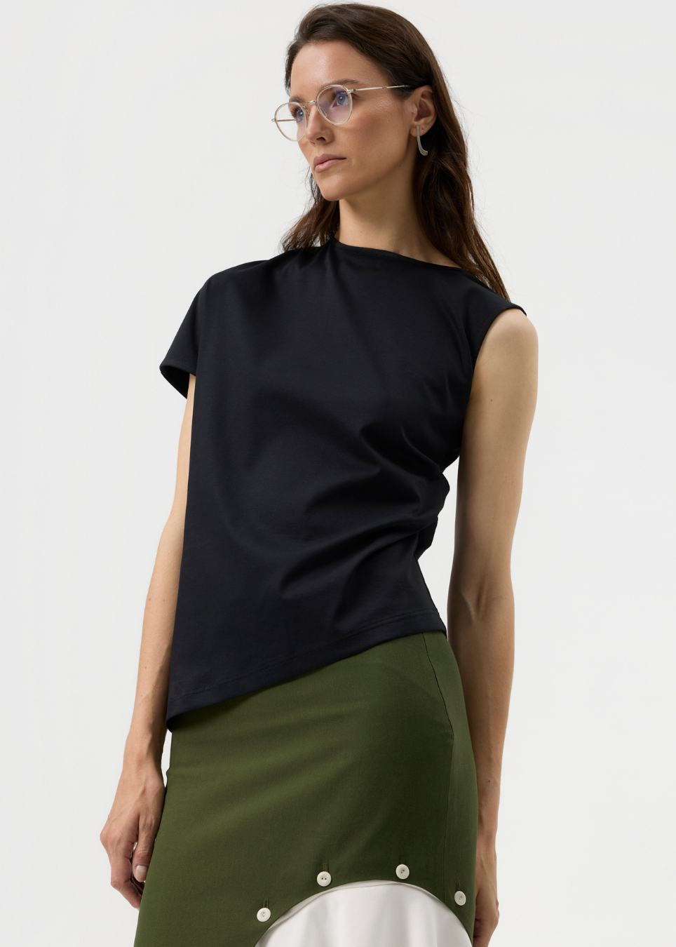 A woman wearing a black short-sleeve top and an olive green skirt with a white panel, photographed against a clean light gray studio background.