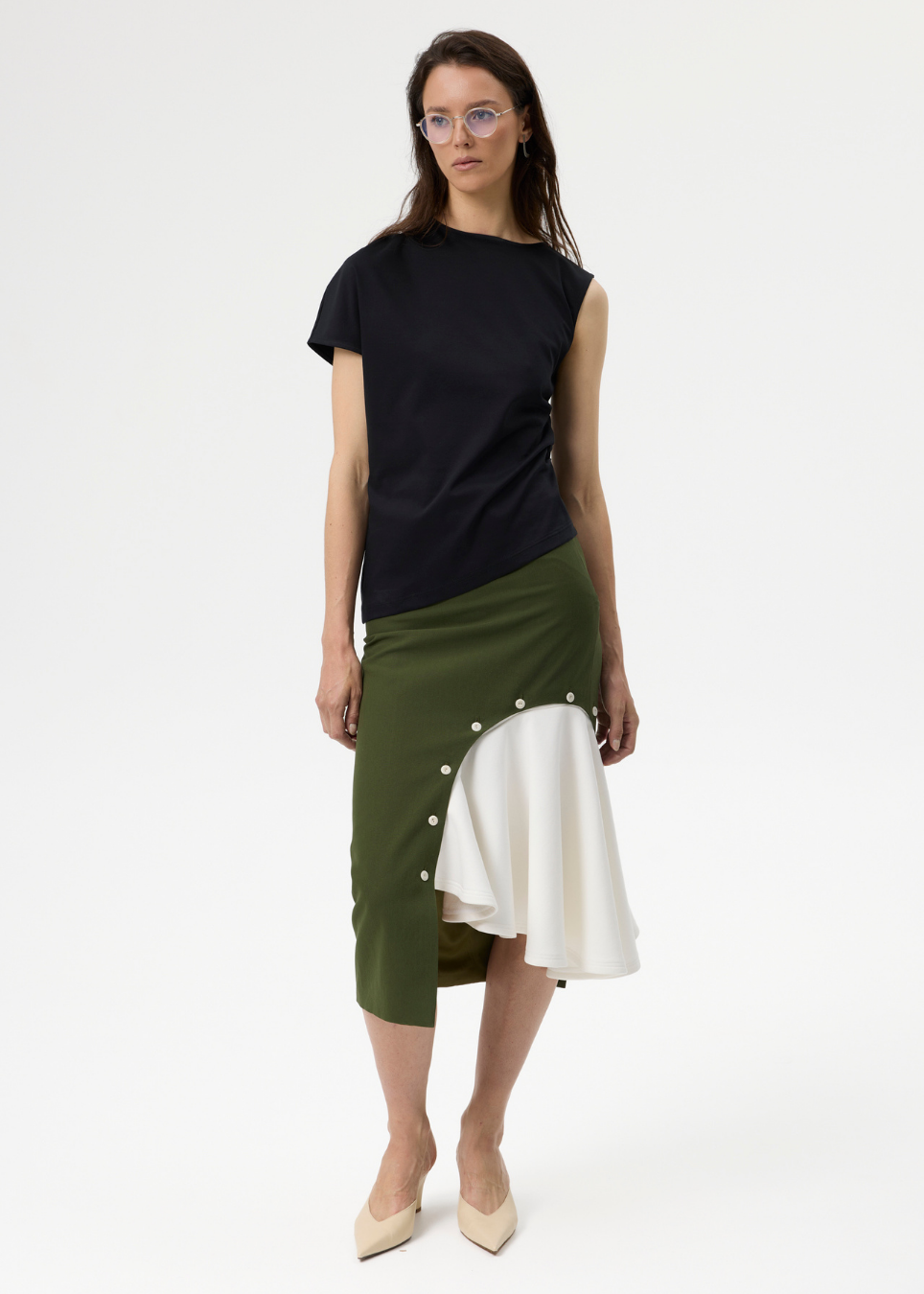 A model stands against a plain white studio background wearing a black short-sleeve top and an asymmetrical olive green and white midi skirt with decorative buttons.