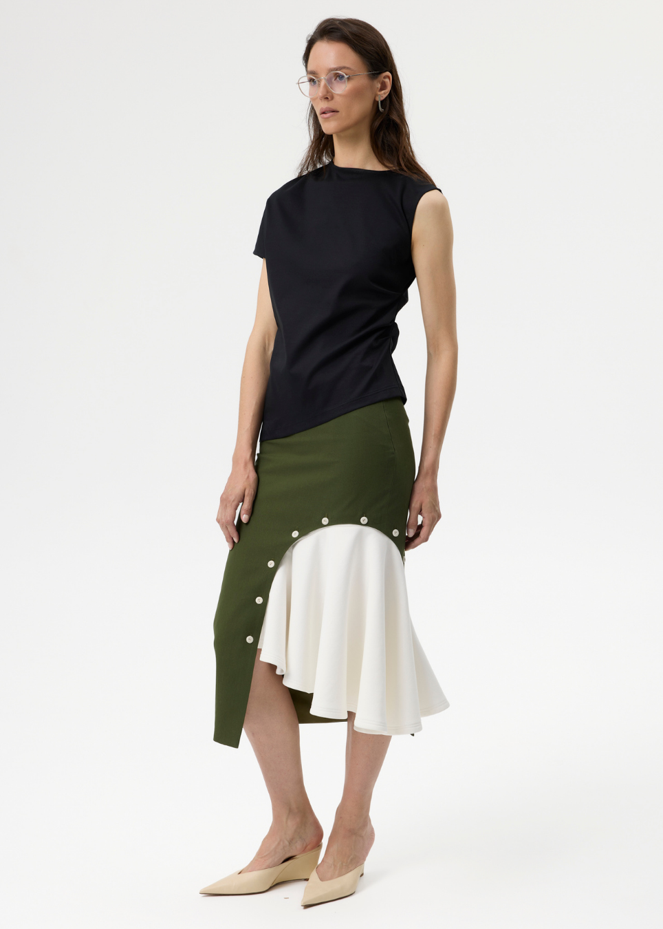 A model wearing a sleeveless black top and a color-block midi skirt in olive green, white, and black, paired with beige pointed-toe flats, against a plain white studio background.