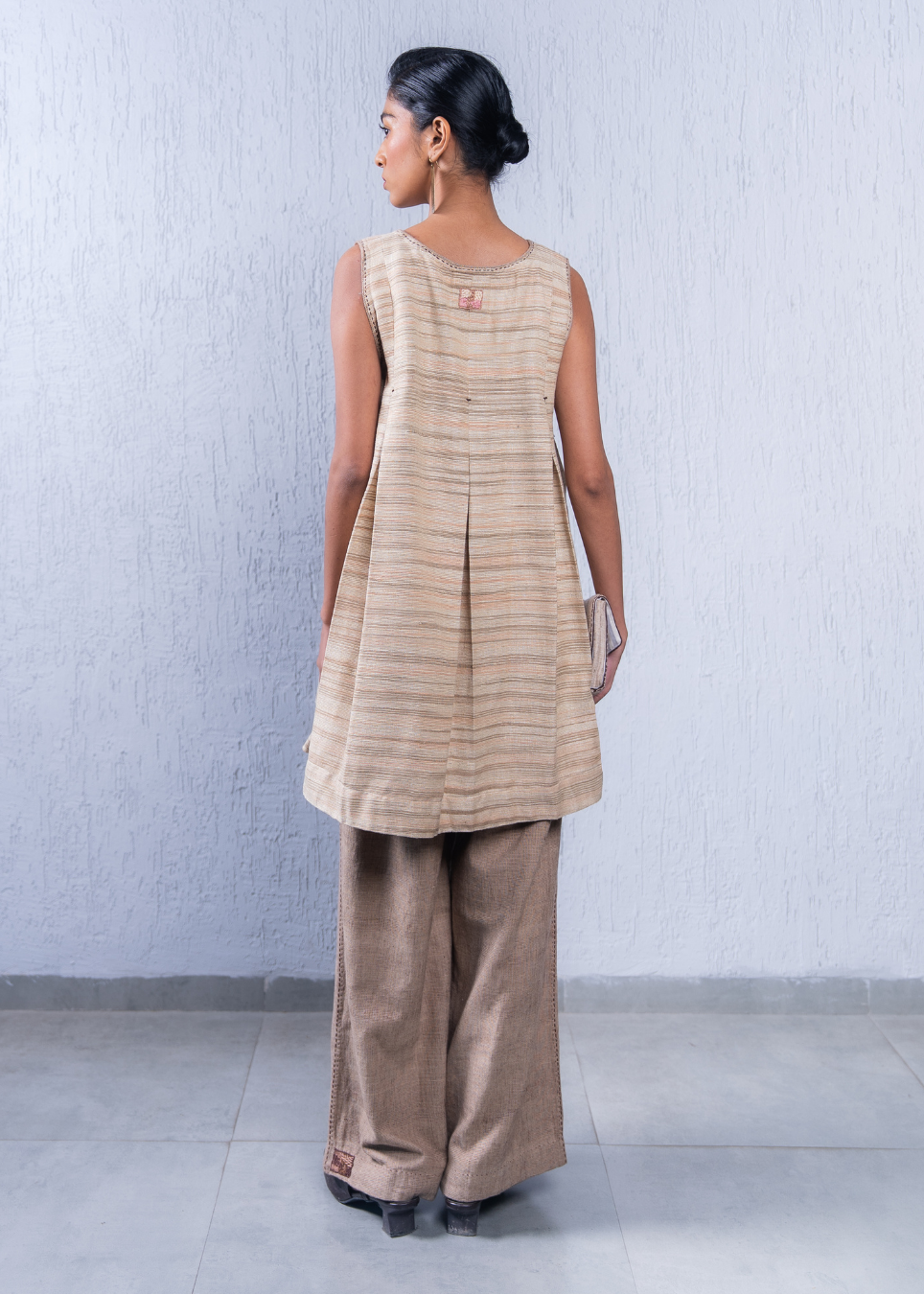 A woman is shown from the back wearing a sleeveless beige and brown striped kurta with wide-leg brown trousers, standing against a textured light gray wall.