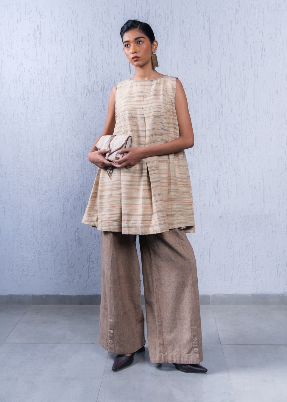 A model stands against a textured light gray wall wearing a sleeveless beige and tan striped top with pleated detailing, wide-leg brown pants, and holding a small beige clutch.