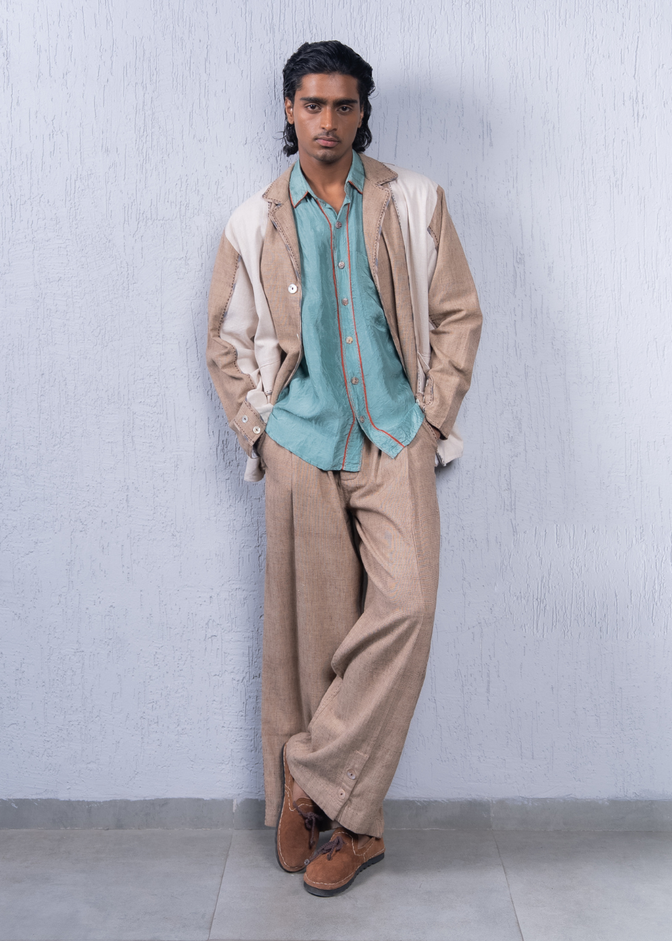 A man stands against a light gray textured wall wearing a beige and off-white jacket, a light blue-green button-up shirt, beige trousers, and brown suede shoes.