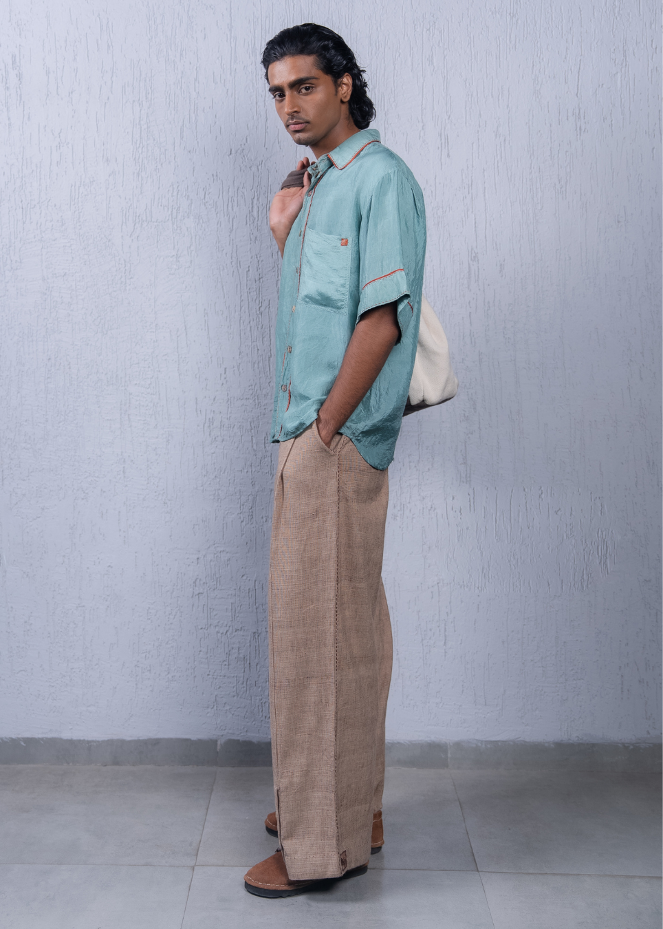 A man wearing a light teal short-sleeve button-up shirt and beige linen trousers, standing against a textured light gray wall.