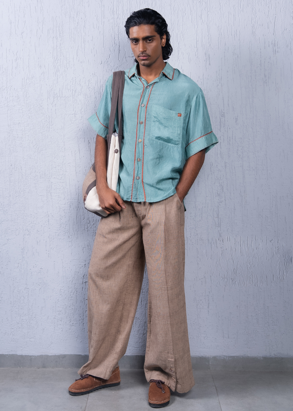 A man stands against a textured light gray wall wearing a light blue-green short-sleeve button-up shirt with orange contrast stitching, beige wide-leg trousers, and brown suede shoes, while carrying a white shoulder bag.