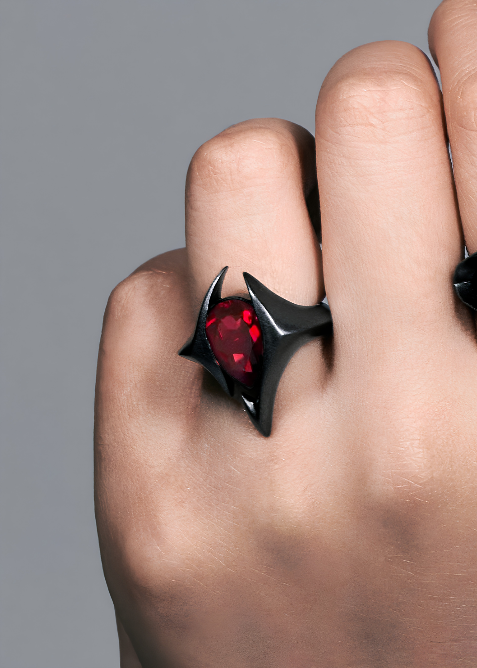 A close-up of a hand wearing a black gothic-style ring with a large, faceted red gemstone.