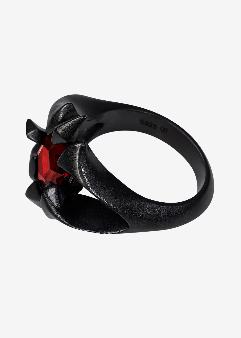 A black metal ring featuring a sculptural, twisted design with a faceted red gemstone at its center, shown on a plain light gray background.