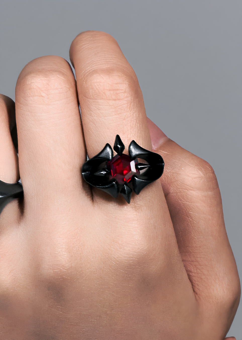 A close-up of a hand wearing a gothic-style black ring with a central faceted red gemstone, set against a neutral gray background.