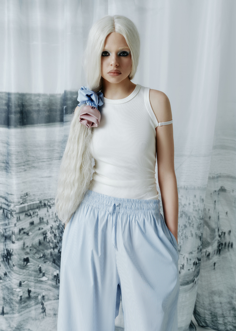 A model with long platinum blonde hair wearing a white ribbed tank top and light blue wide-leg pants, standing in front of a white curtain and a black and white landscape print.