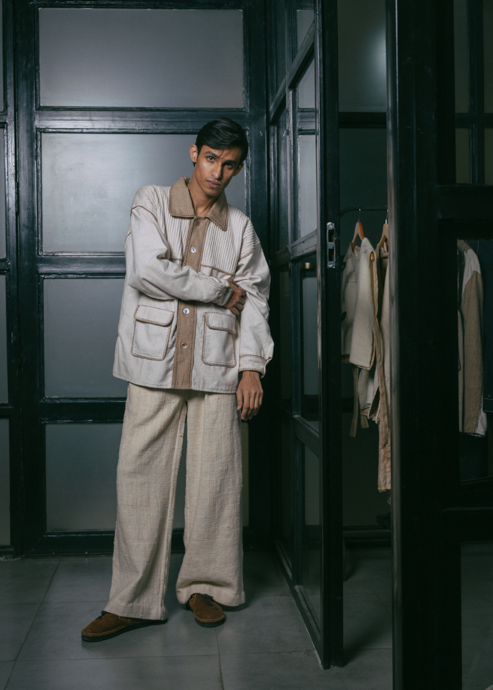A man wearing a light beige two-piece lounge set consisting of a long-sleeve button-up shirt and matching wide-leg trousers, standing in a dark doorway.