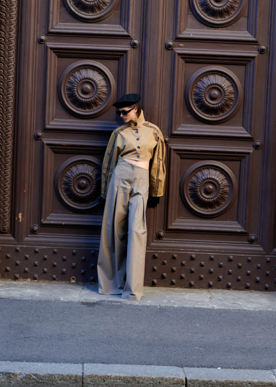A model wearing a beige military-style jacket and matching wide-leg trousers, a black cap, and sunglasses, posing in front of a large, dark brown decorative wooden door.