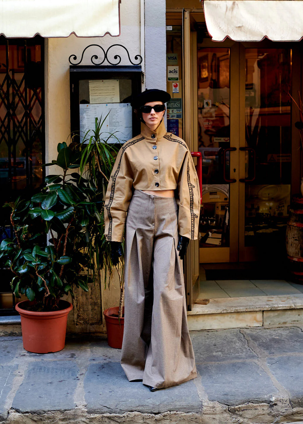 A model stands on a stone sidewalk in front of a shop entrance, wearing a beige military-style cropped jacket with black trim, matching wide-leg trousers, black gloves, and a black beret.