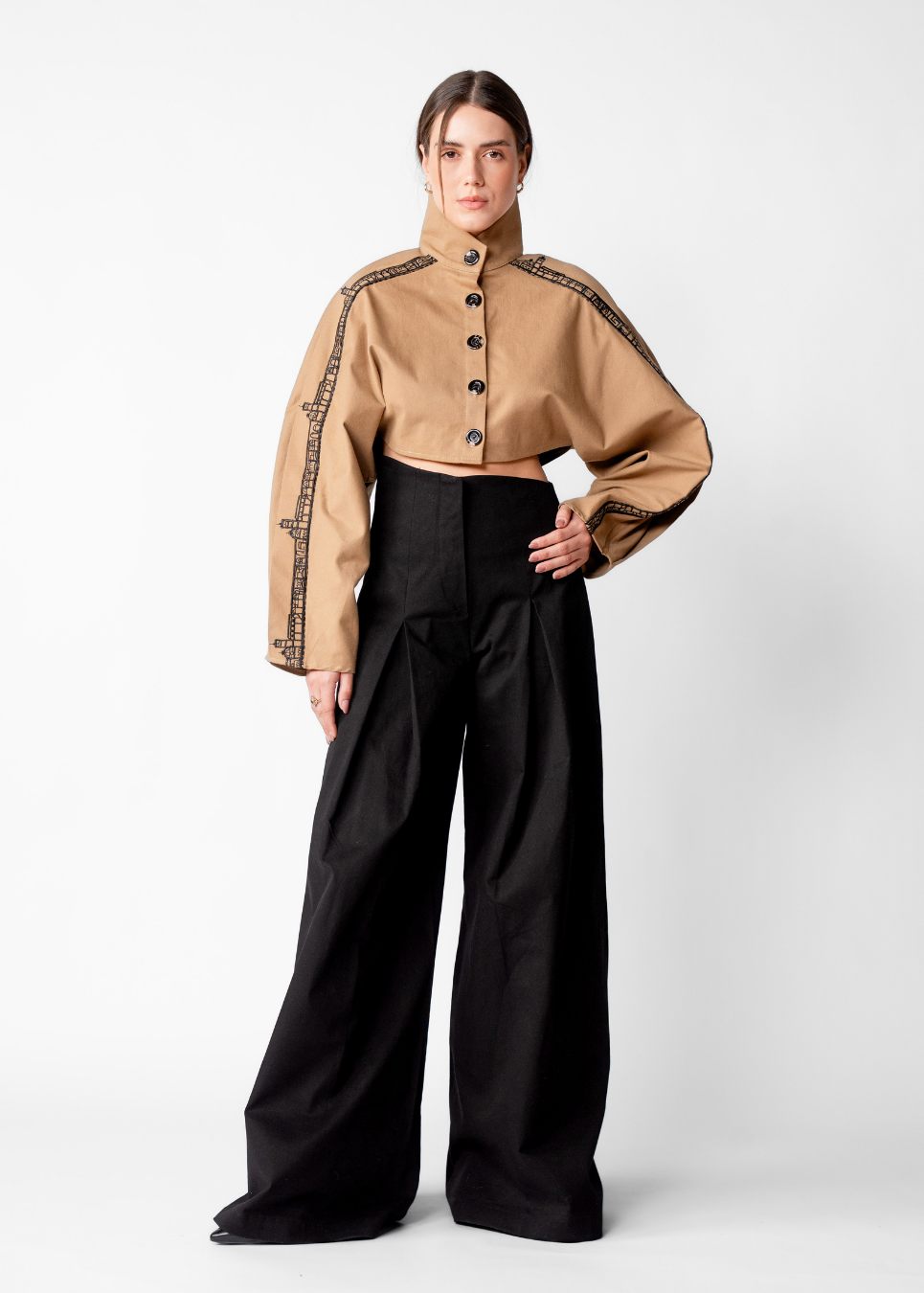 A model stands against a light gray studio background wearing a cropped tan jacket with black embroidered trim and black wide-leg trousers.