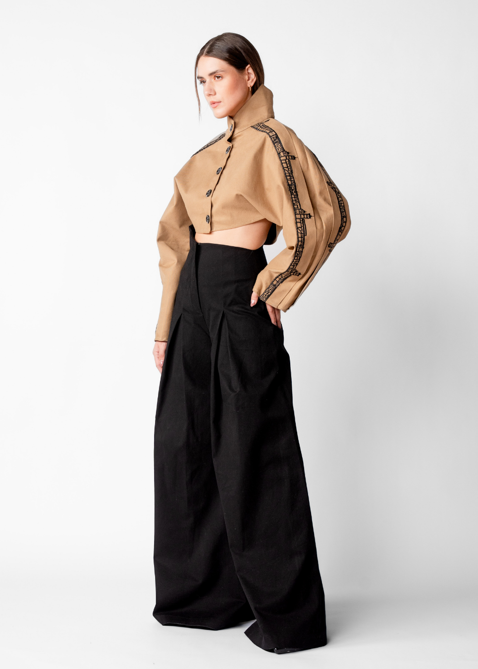 A model wearing a tan cropped button-up shirt with black patterned trim on the sleeves and black wide-leg trousers, standing against a light gray studio background.