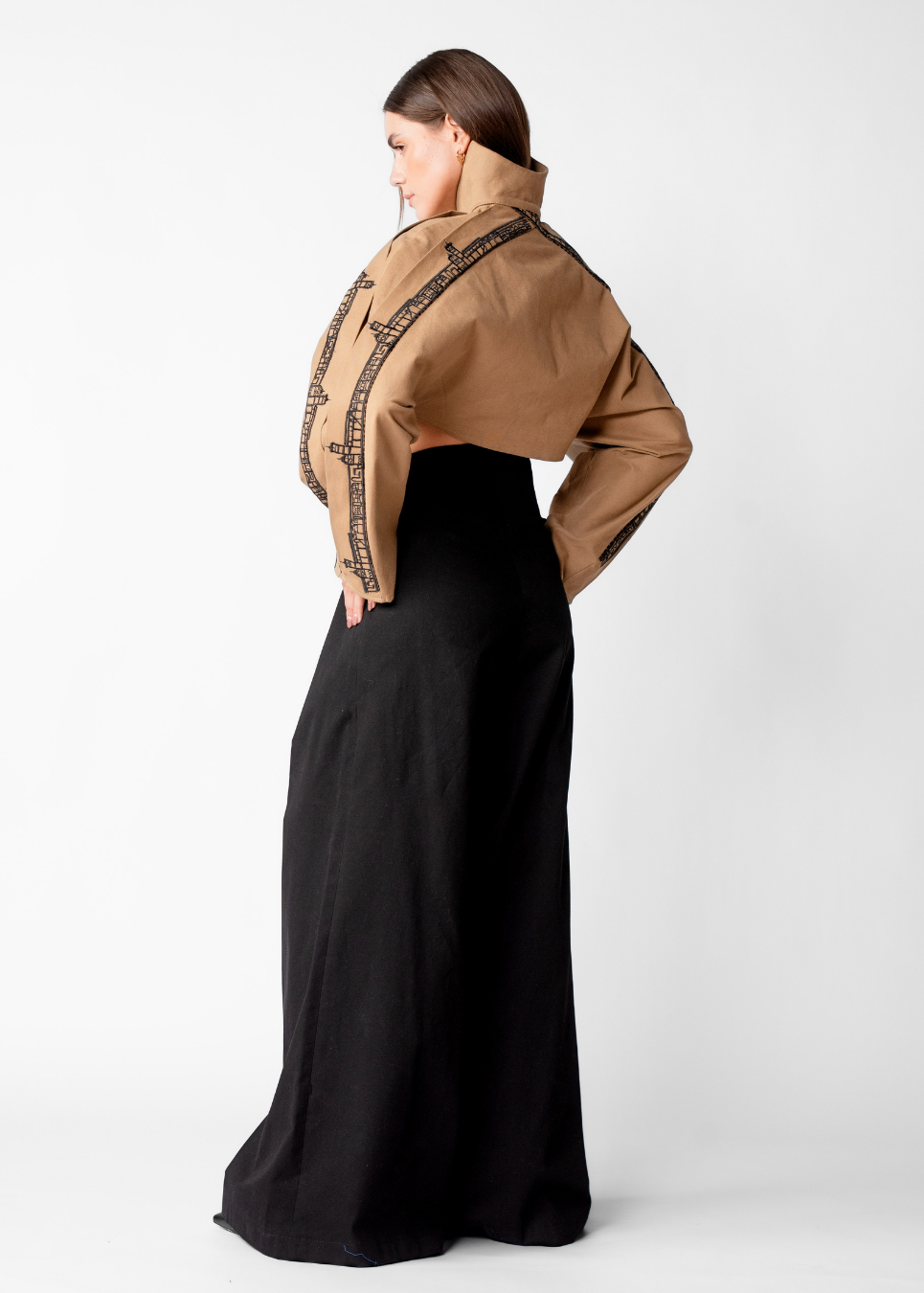 A woman shown from the back wearing a tan cropped jacket with black geometric embroidery and a long black skirt, photographed against a light gray studio background.