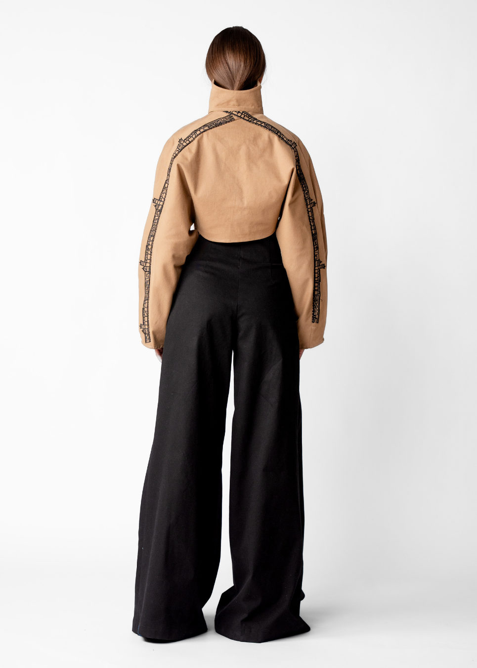 A model shown from the back wearing a tan cropped jacket with black chain-link embroidery down the sleeves and black wide-leg trousers, set against a plain white studio background.