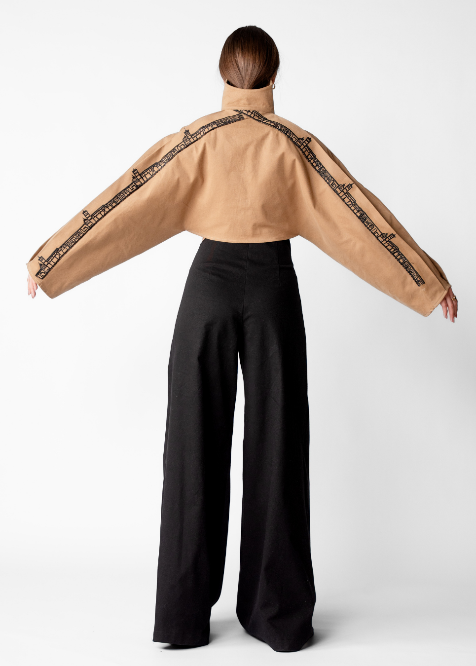 A model shown from the back wearing a cropped tan jacket with black patterned trim along the shoulders and sleeves, paired with black wide-leg trousers, against a light gray studio background.