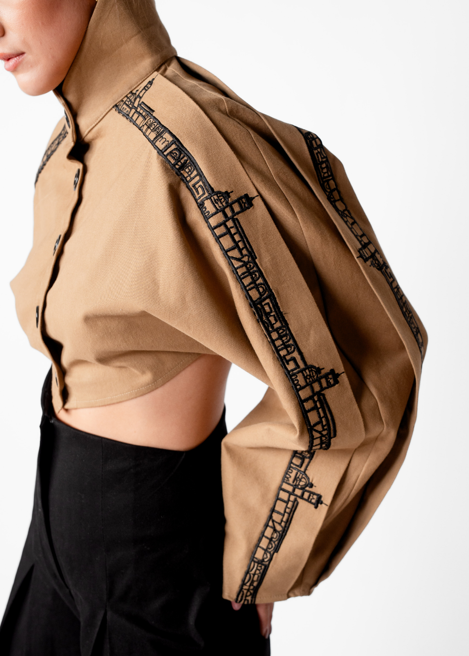 A close-up of a model wearing a cropped tan long-sleeve shirt with dramatic puffed sleeves and black architectural line-art embroidery down the sleeves, paired with a black skirt.