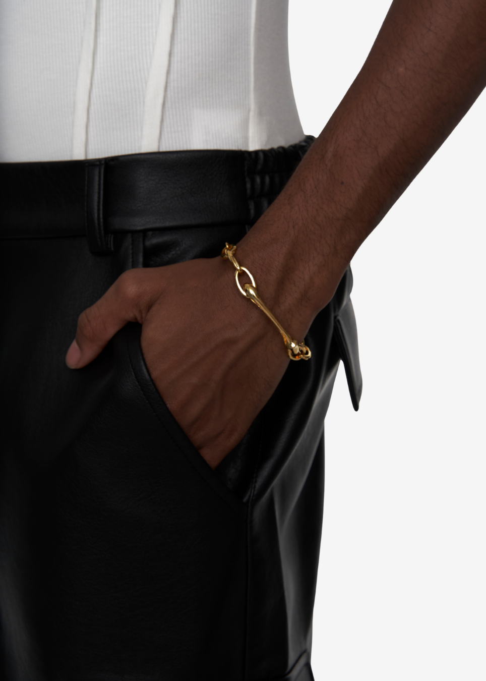 A close-up of a person wearing a gold chain bracelet with a knotted loop detail on the wrist, styled with black leather trousers and a white top against a light gray background.