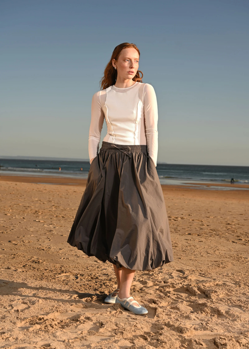 A woman stands on a sandy beach wearing a white long-sleeve mock-neck top and a dark gray pleated midi skirt, paired with light blue shoes.