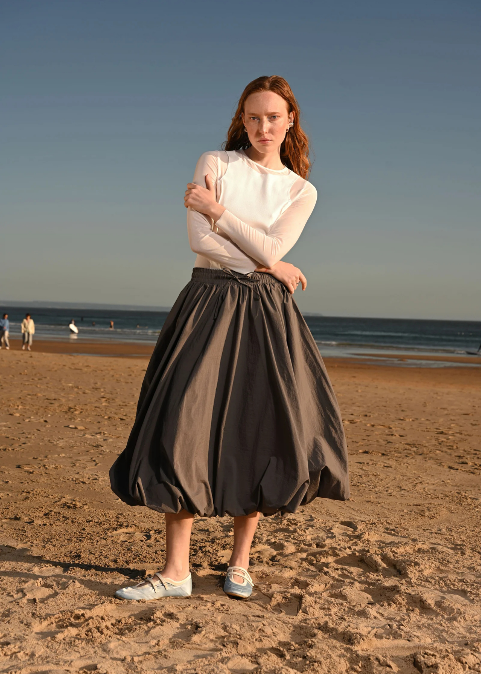 A woman stands on a sandy beach wearing a white long-sleeve top and a dark gray pleated midi skirt, paired with white sneakers. The background shows the ocean and a clear blue sky.