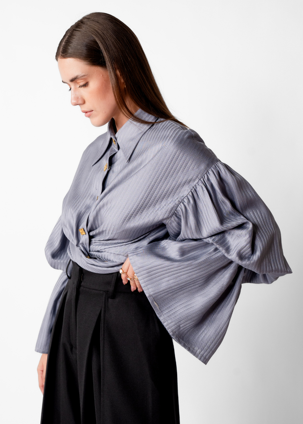 A woman wearing a gray ribbed blouse with dramatic ruffled bell sleeves and a tied waist, paired with black trousers, photographed against a plain light gray studio background.