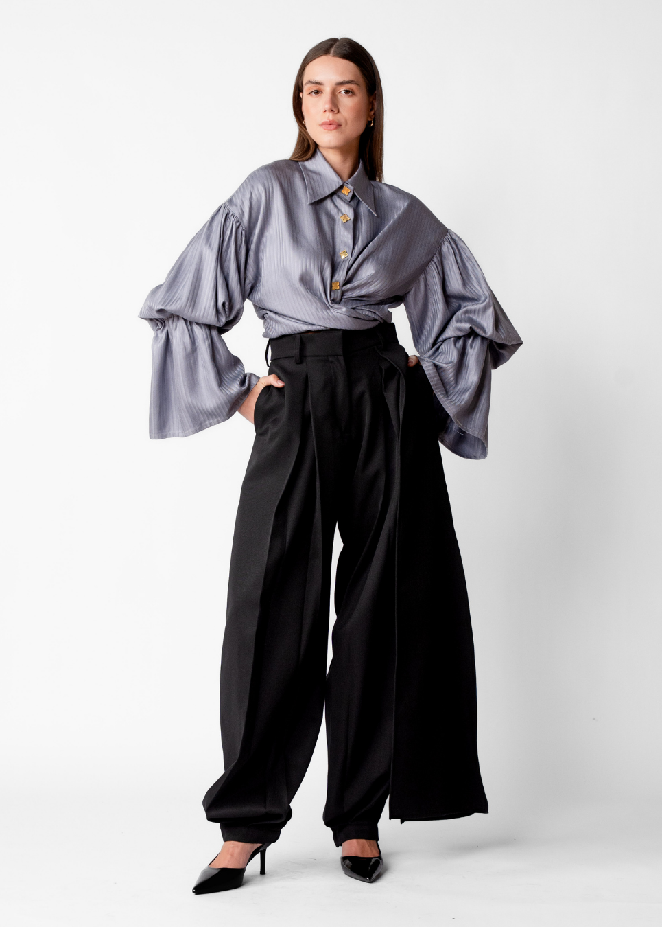 A model stands against a light gray studio background wearing a lavender-gray satin blouse with dramatic puffed sleeves and a buttoned collar, paired with black wide-leg trousers and black pointed heels.