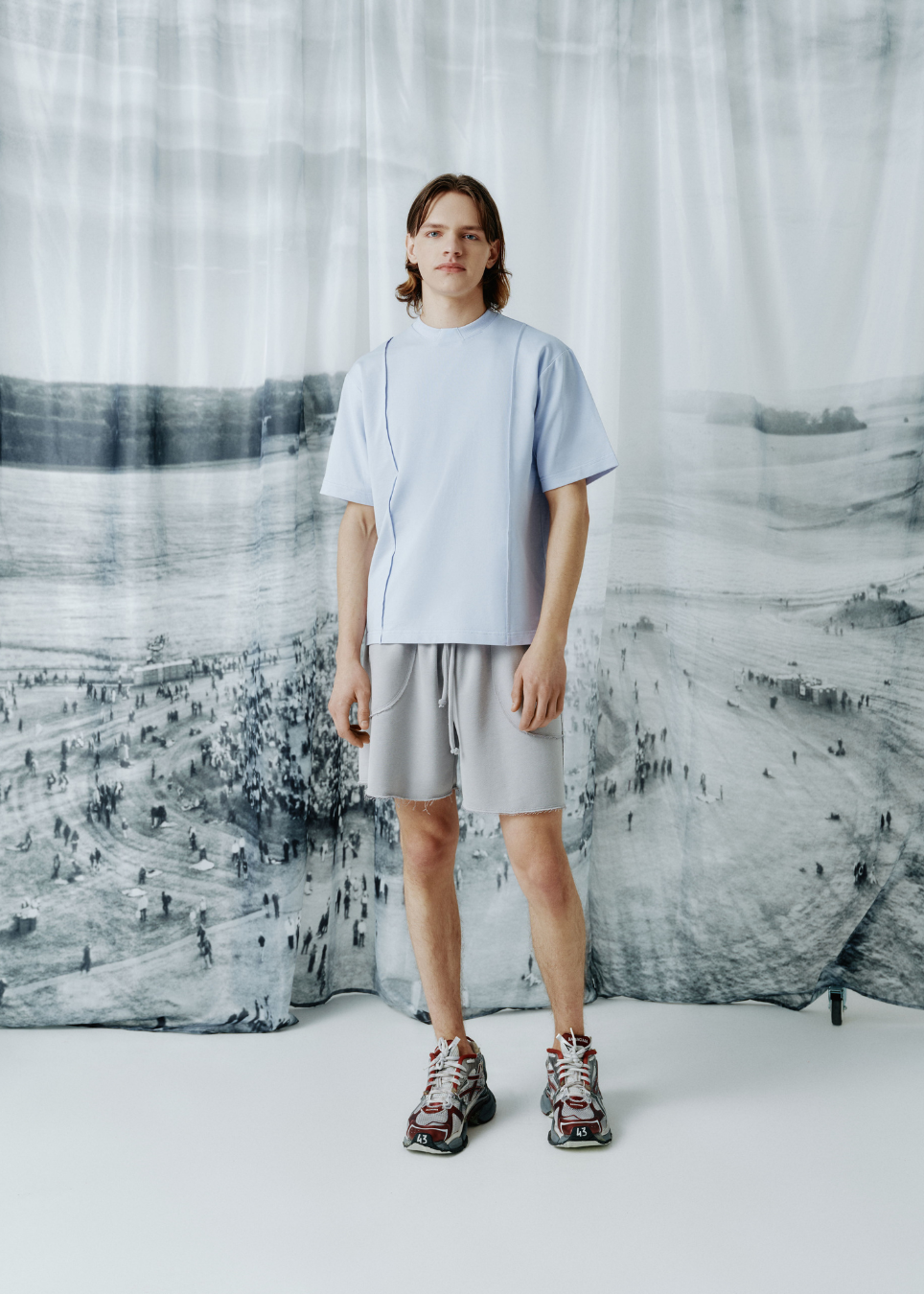 A model stands in a studio wearing a light blue oversized short-sleeve top, light gray drawstring shorts, and patterned sneakers, posed in front of a large black-and-white landscape photograph backdrop.