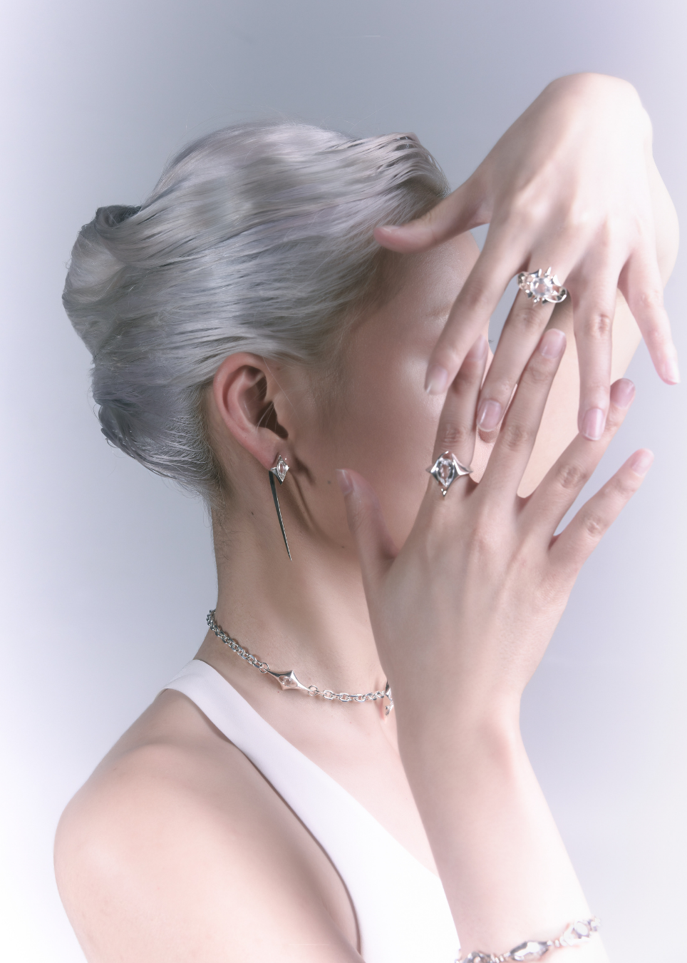 A woman with platinum blonde hair shown in profile wearing delicate silver jewelry, including a long drop earring, rings, and a bracelet, against a light gray studio background.