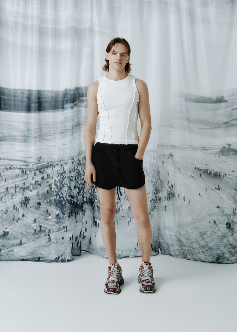 A model stands in front of a large black-and-white scenic print backdrop, wearing a sleeveless white ribbed top, black shorts, and patterned sneakers.