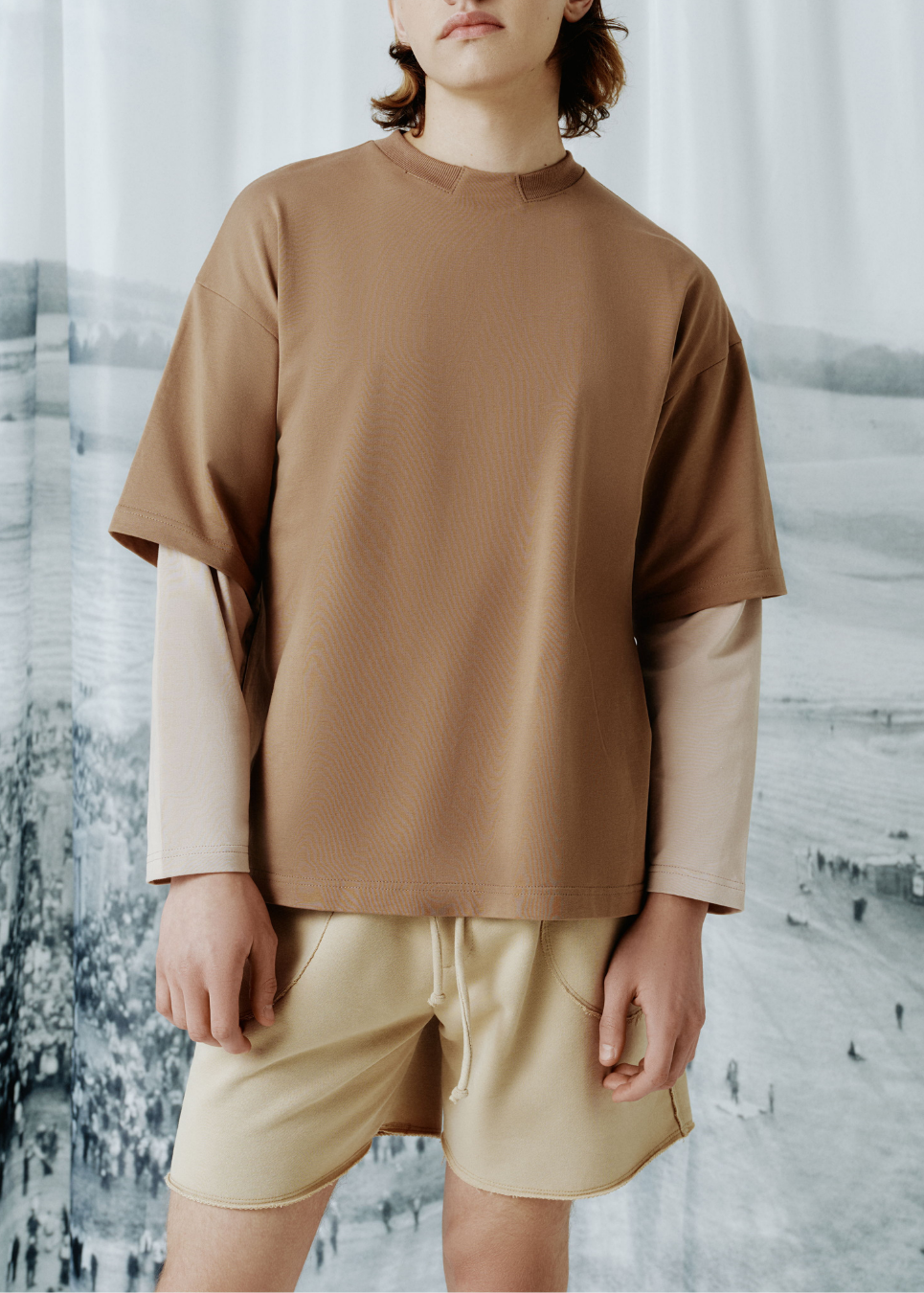 A model wearing a brown ribbed short-sleeve t-shirt layered over a beige long-sleeve shirt, paired with beige shorts, standing in front of a scenic landscape backdrop.