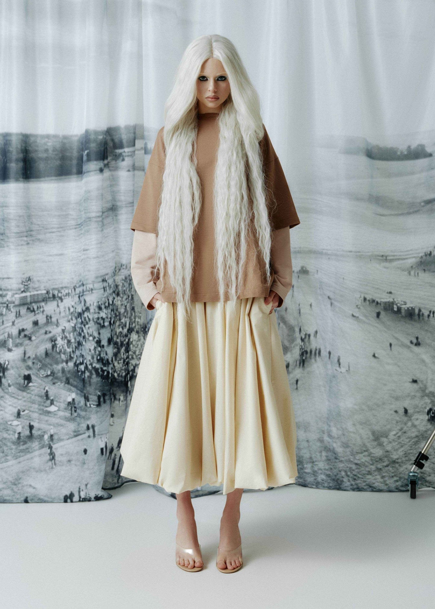 A model with long, straight white hair stands in front of a large black-and-white scenic backdrop, wearing a layered outfit consisting of a brown short-sleeve top over a light beige long-sleeve shirt, a cream pleated midi skirt, and nude high-heeled sandals.