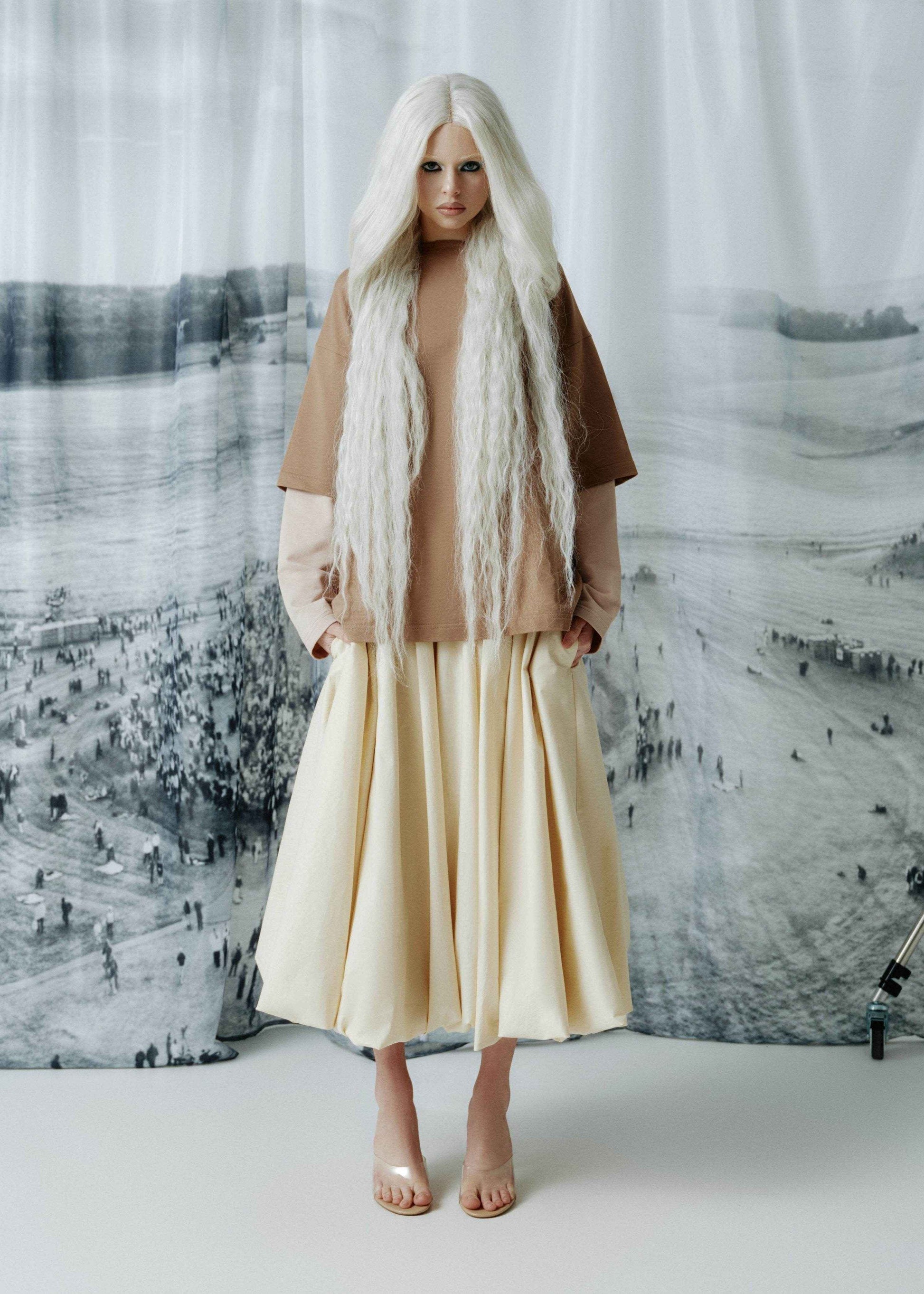 A model with long, straight white hair stands in front of a large black-and-white scenic backdrop, wearing a layered outfit consisting of a brown short-sleeve top over a light beige long-sleeve shirt, a cream pleated midi skirt, and nude high-heeled sandals.