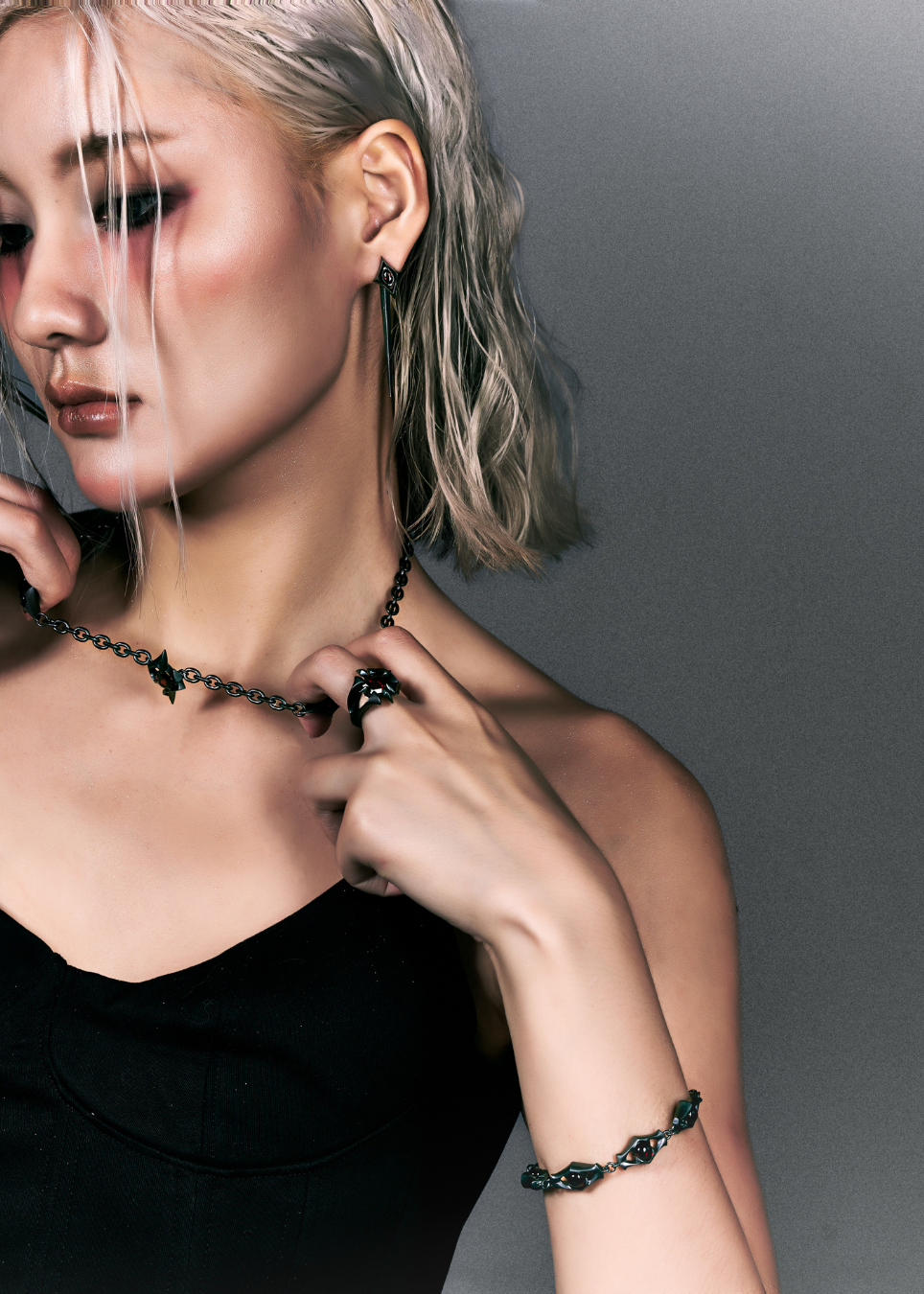 A close-up lifestyle shot of a model with platinum blonde hair wearing a black chain necklace with a central black heart pendant, a matching black beaded bracelet, and a black ring against a dark gray background.