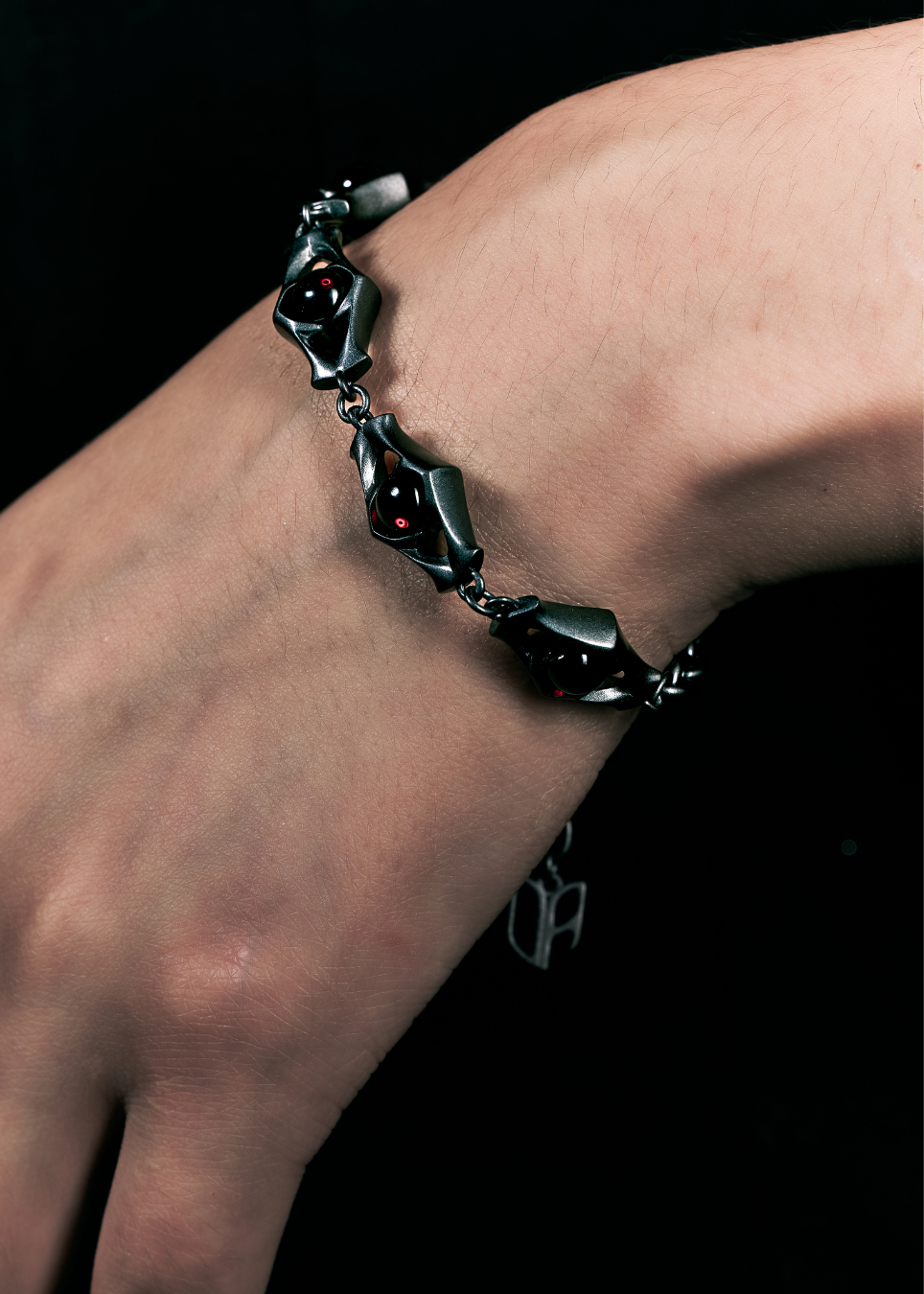 A close-up of a hand wearing a dark metallic bracelet with red gemstone accents against a black background.