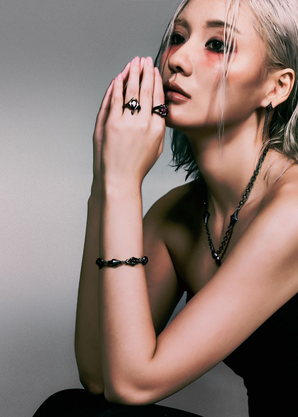 A model with platinum blonde hair and dark eye makeup poses with her hands near her face, showcasing black gothic jewelry including a beaded bracelet, a chain necklace, and black rings with dark stones.
