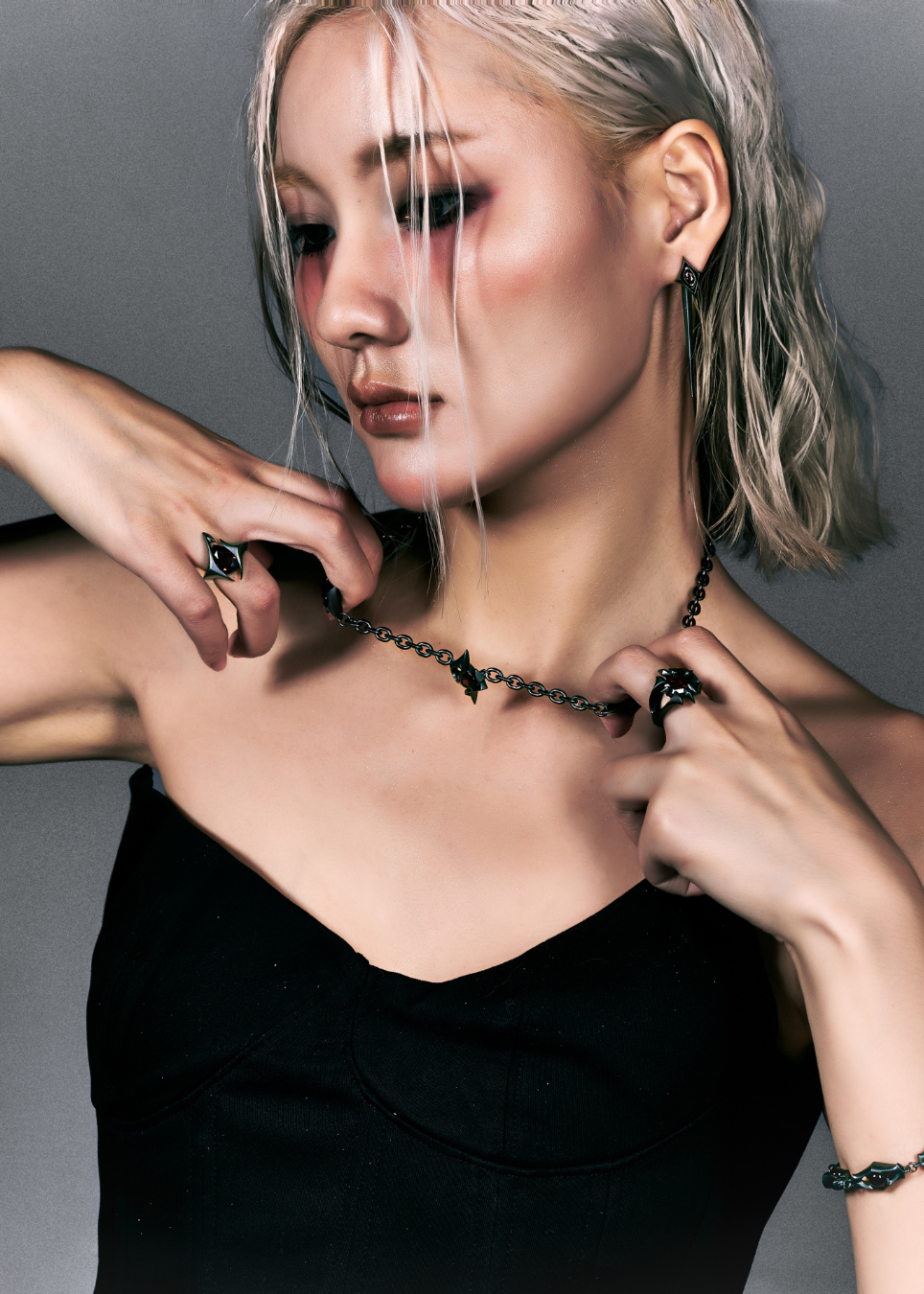 A close-up portrait of a woman with platinum blonde hair wearing a black chain necklace with a small heart pendant, a black top, and black rings, posed against a dark gray background.