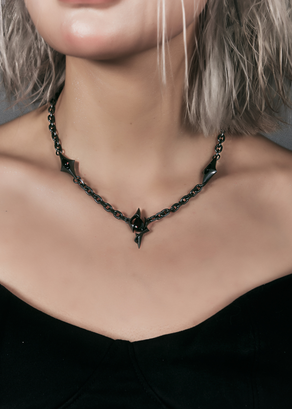A close-up of a model wearing a dark metallic gothic chain necklace with sharp star and diamond-shaped spikes, paired with a black top.