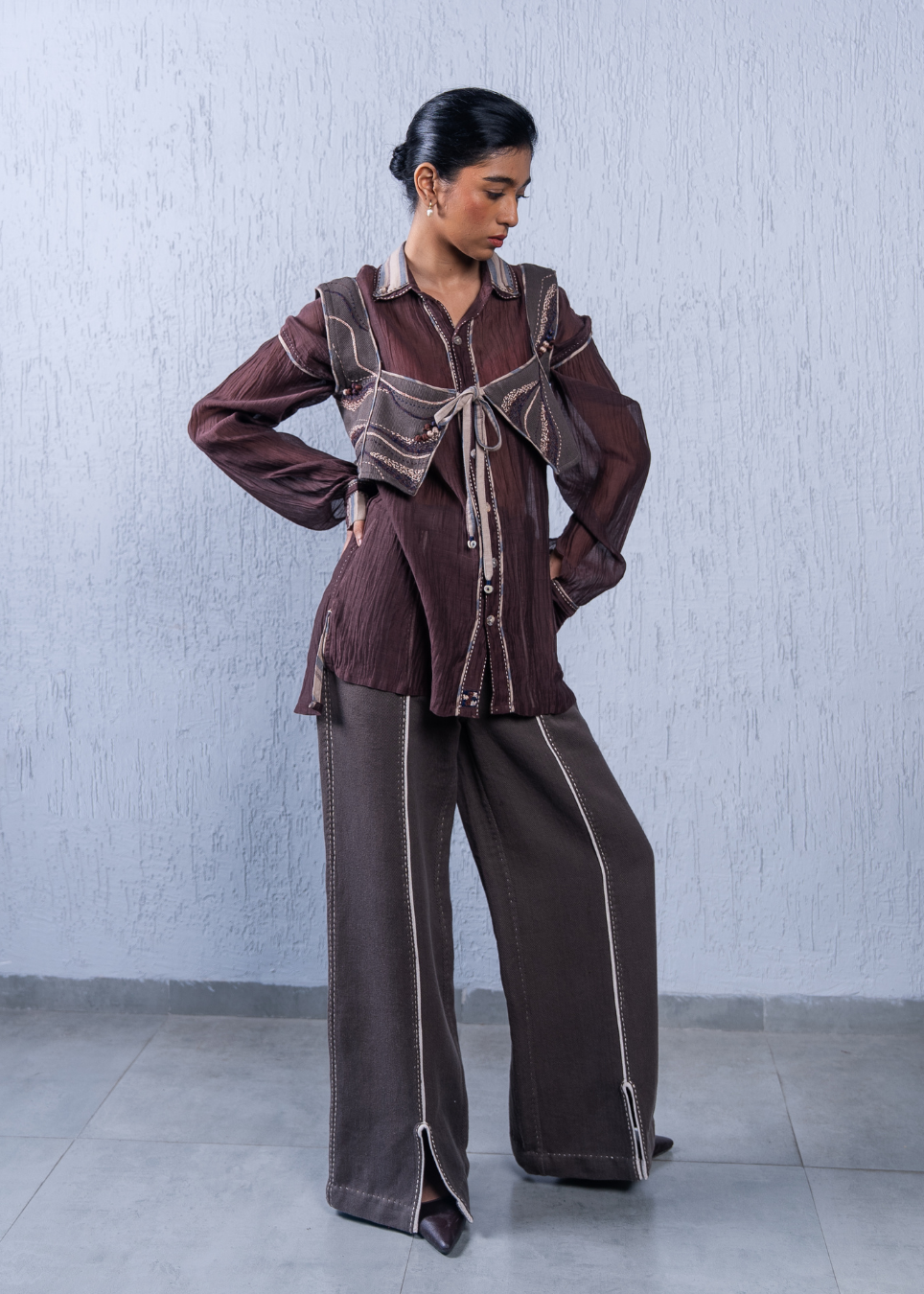 A model wearing a brown and beige two-piece outfit consisting of a long-sleeve button-up shirt with tie details and matching wide-leg trousers with contrast stitching, standing against a textured light gray wall.