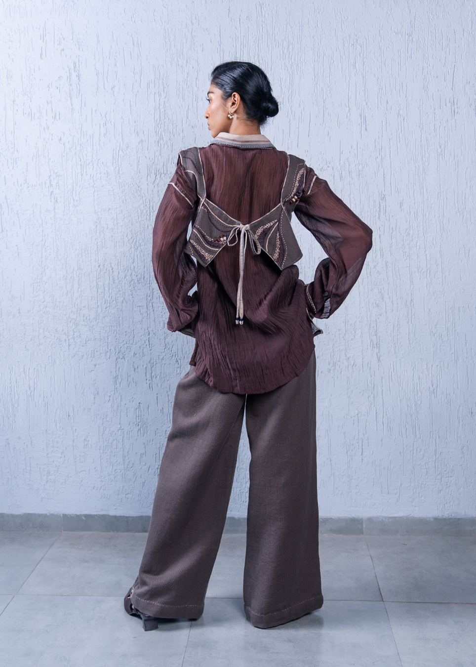 A model is shown from the back wearing a brown long-sleeve blouse with a unique tie-back design and matching wide-leg trousers, set against a textured light gray wall.