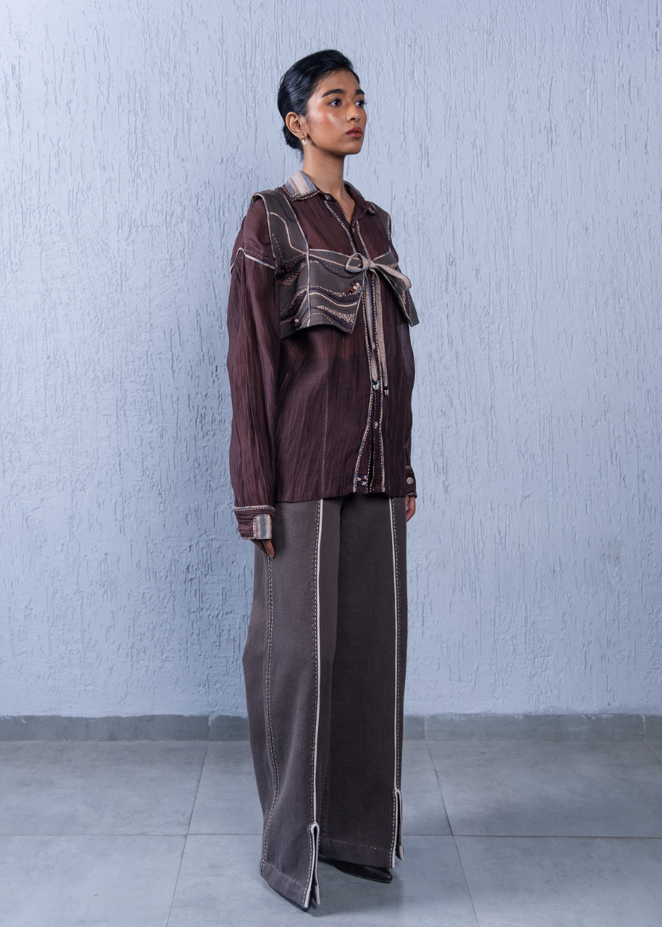 A model stands in profile wearing a dark brown long-sleeve button-up shirt with a large tie detail at the collar and matching wide-leg trousers, set against a textured light gray wall.