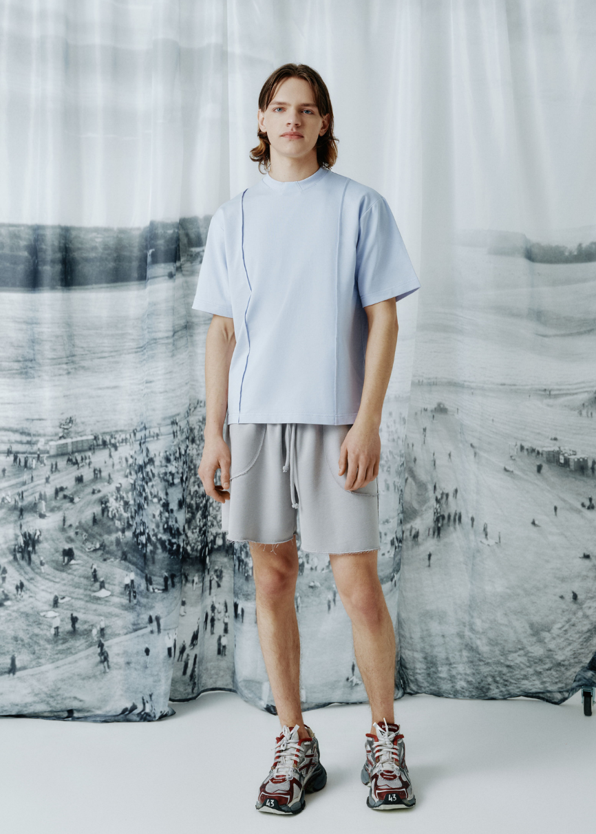 A model stands in a studio wearing a light blue t-shirt, light gray shorts, and patterned sneakers, posed in front of a large black and white scenic backdrop.